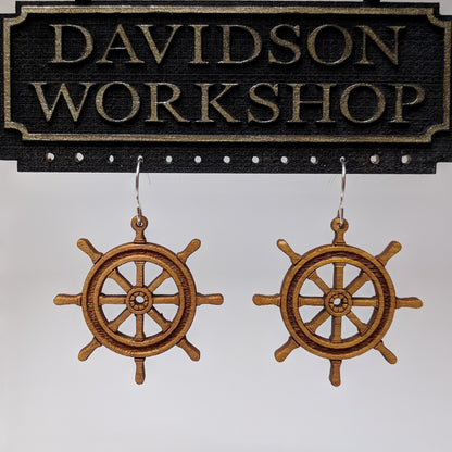 Pair of wooden earrings with silver stainless steel hooks. They are brown pirate ship wheels with 8 handles and a spoked center. Made from birch wood hanging from a model Davidson Workshop sign against a white background. (Close up View)