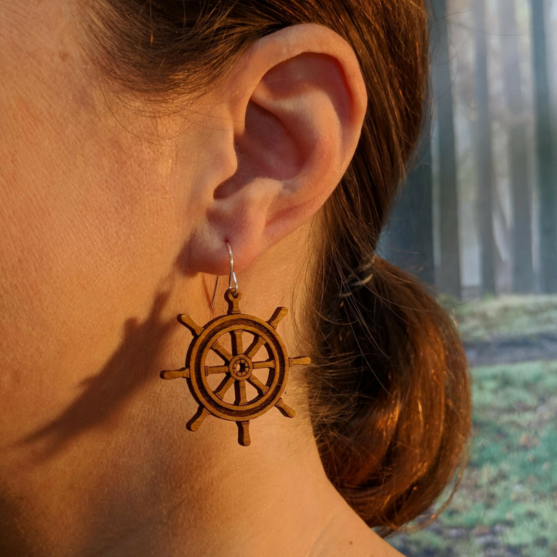 Pair of wooden earrings with silver stainless steel hooks. They are brown pirate ship wheels with 8 handles and a spoked center. Made from birch wood, being modeled by a women in front of a nature scene.