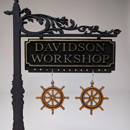 Pair of wooden earrings with silver stainless steel hooks. They are brown pirate ship wheels with 8 handles and a spoked center. Made from birch wood hanging from a model Davidson Workshop sign against a white background. 