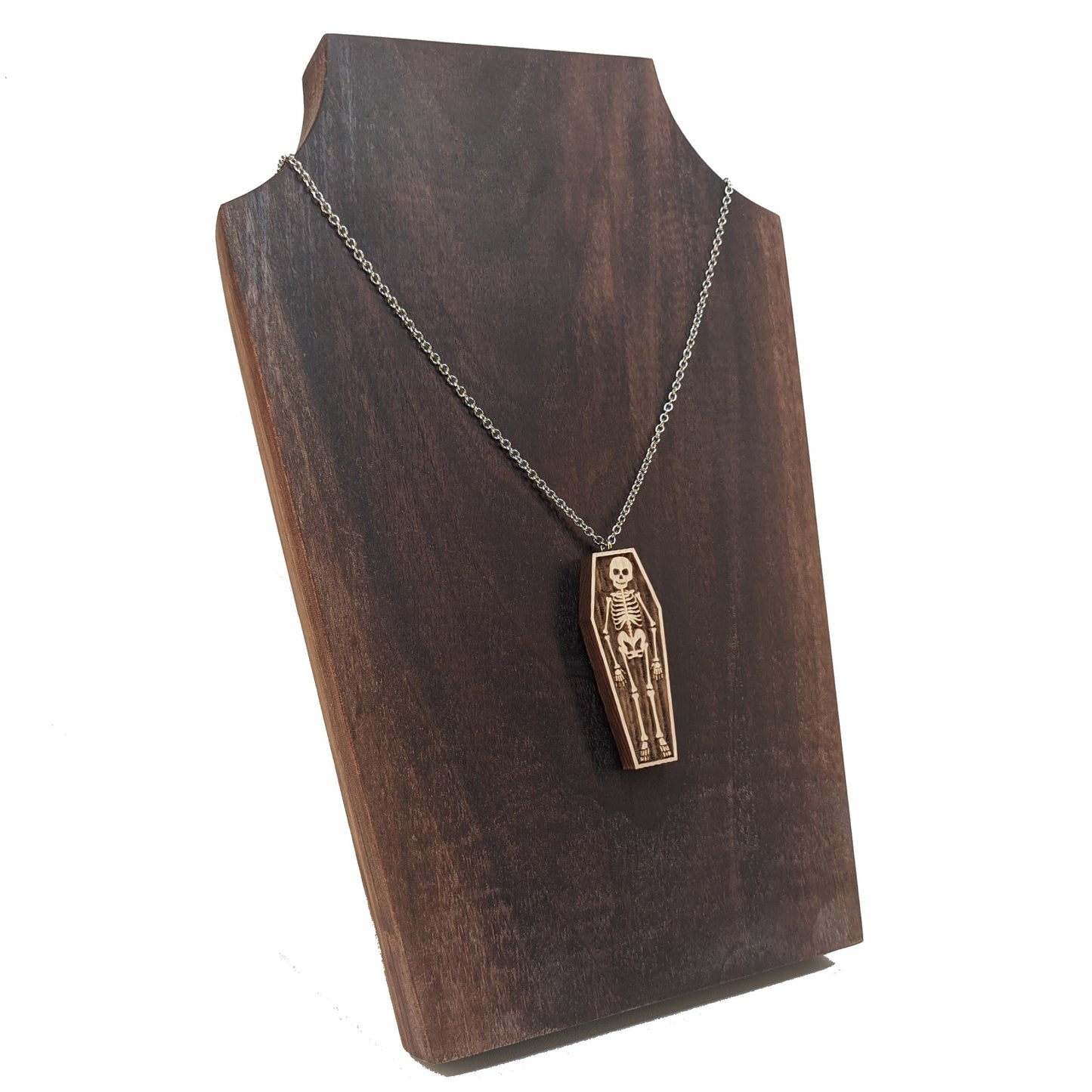 Wooden necklace pendant created in the shape a skeleton lying in a coffin. Hanging from a silver stainless steel chain against a  dark walnut background.