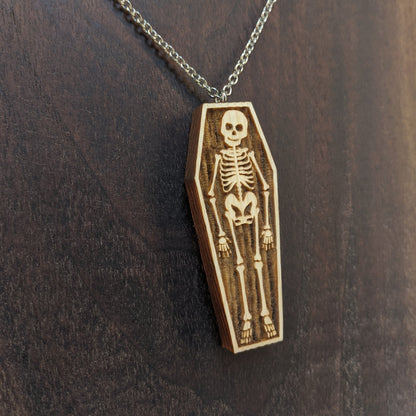 Wooden necklace pendant created in the shape a skeleton lying in a coffin. Hanging from a silver stainless steel chain against a  dark walnut background.