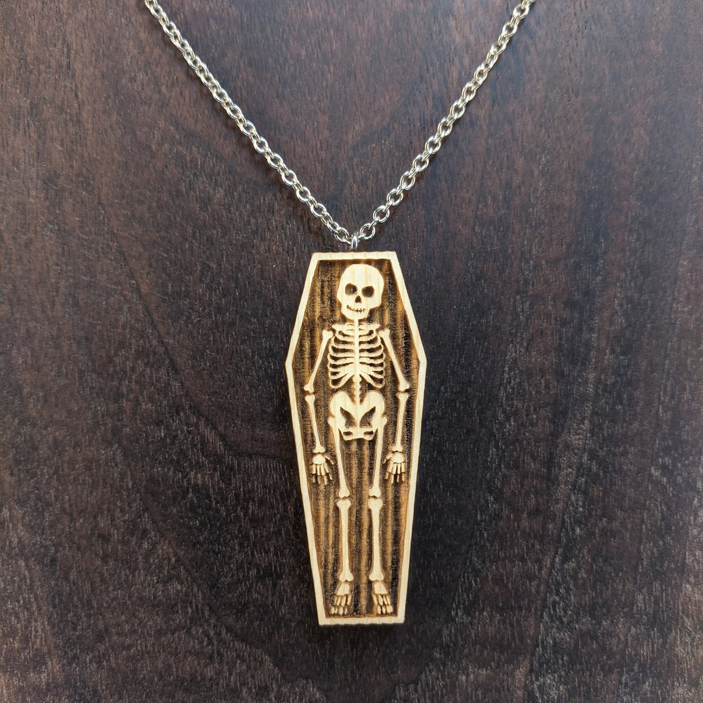 Wooden necklace pendant created in the shape a skeleton lying in a coffin. Hanging from a silver stainless steel chain against a  dark walnut background.