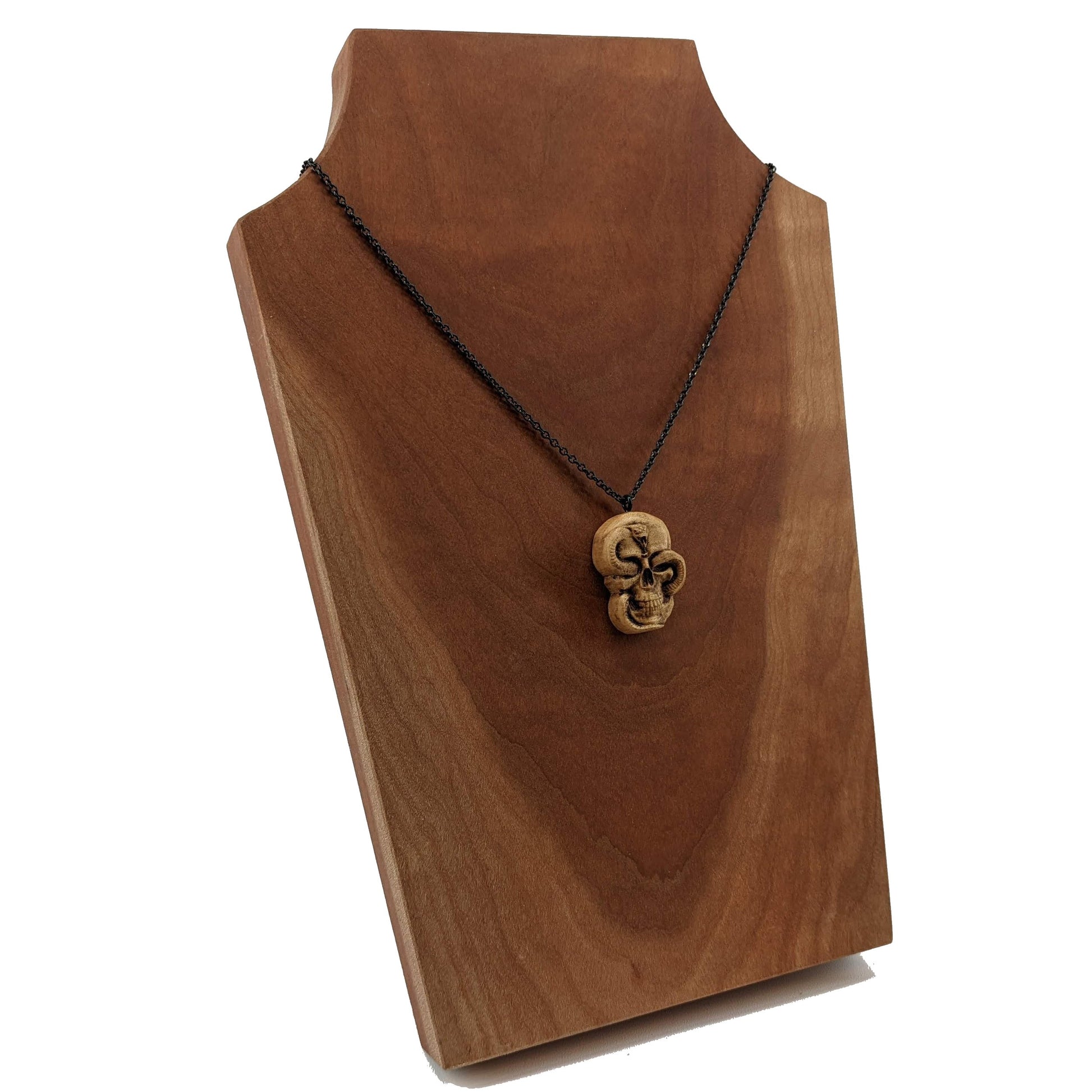 Wooden necklace pendant carved in the shape of a skull with a snake crawling through the eyes. Made from hard maple wood and hanging from a black stainless steel chain against a cherry wood display.