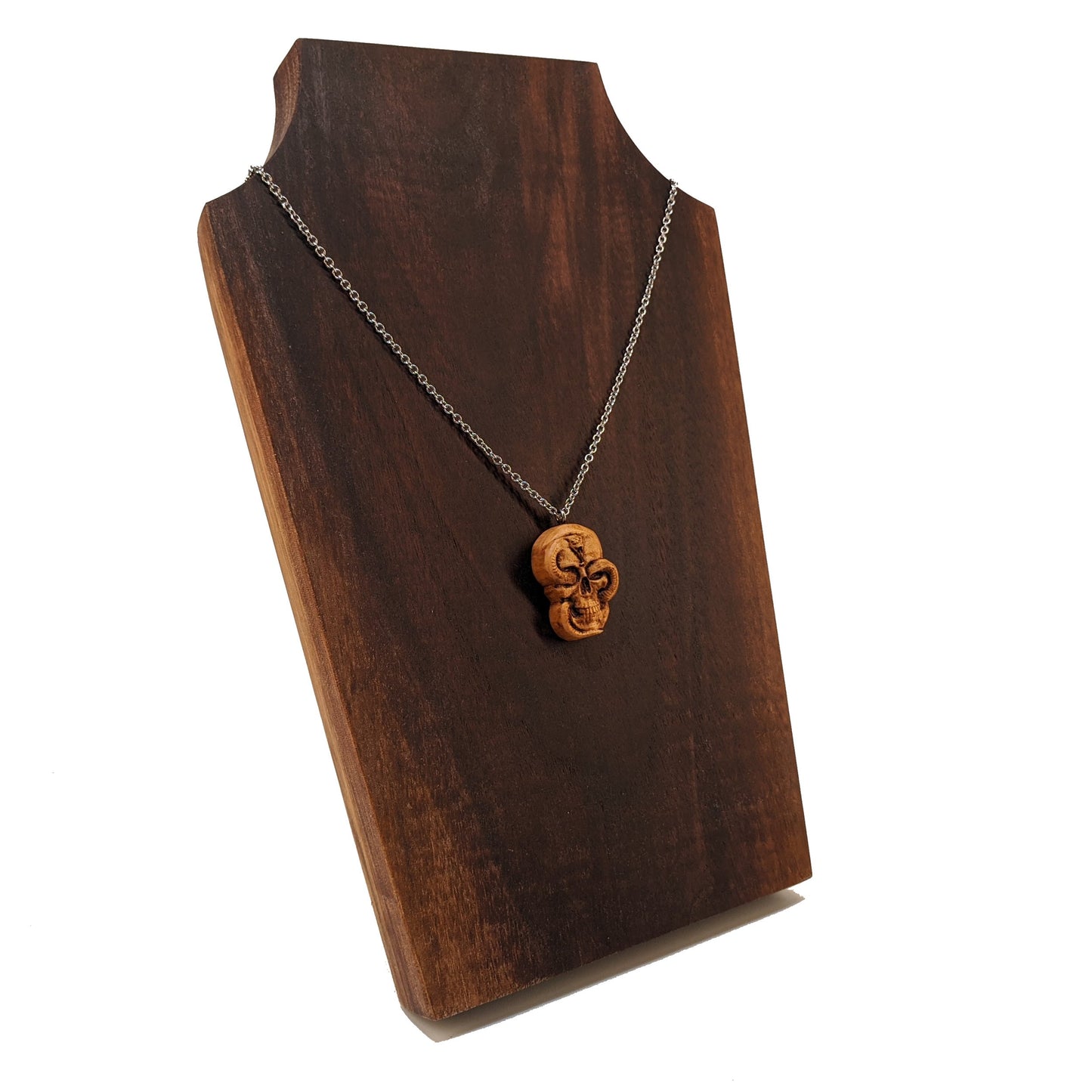 Wooden necklace pendant carved in the shape of a skull with a snake crawling through the eyes. Made from hard maple wood and hanging from a silver stainless steel chain against a dark walnut wood display.