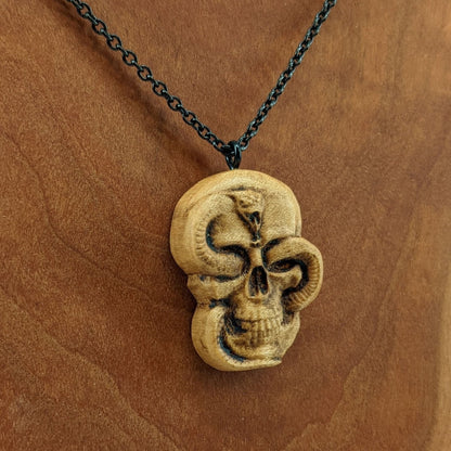 Wooden necklace pendant carved in the shape of a skull with a snake crawling through the eyes. Made from hard maple wood and hanging from a black stainless steel chain against a cherry wood display.