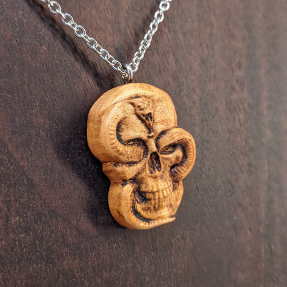 Wooden necklace pendant carved in the shape of a skull with a snake crawling through the eyes. Made from hard maple wood and hanging from a silver stainless steel chain against a dark walnut wood background.