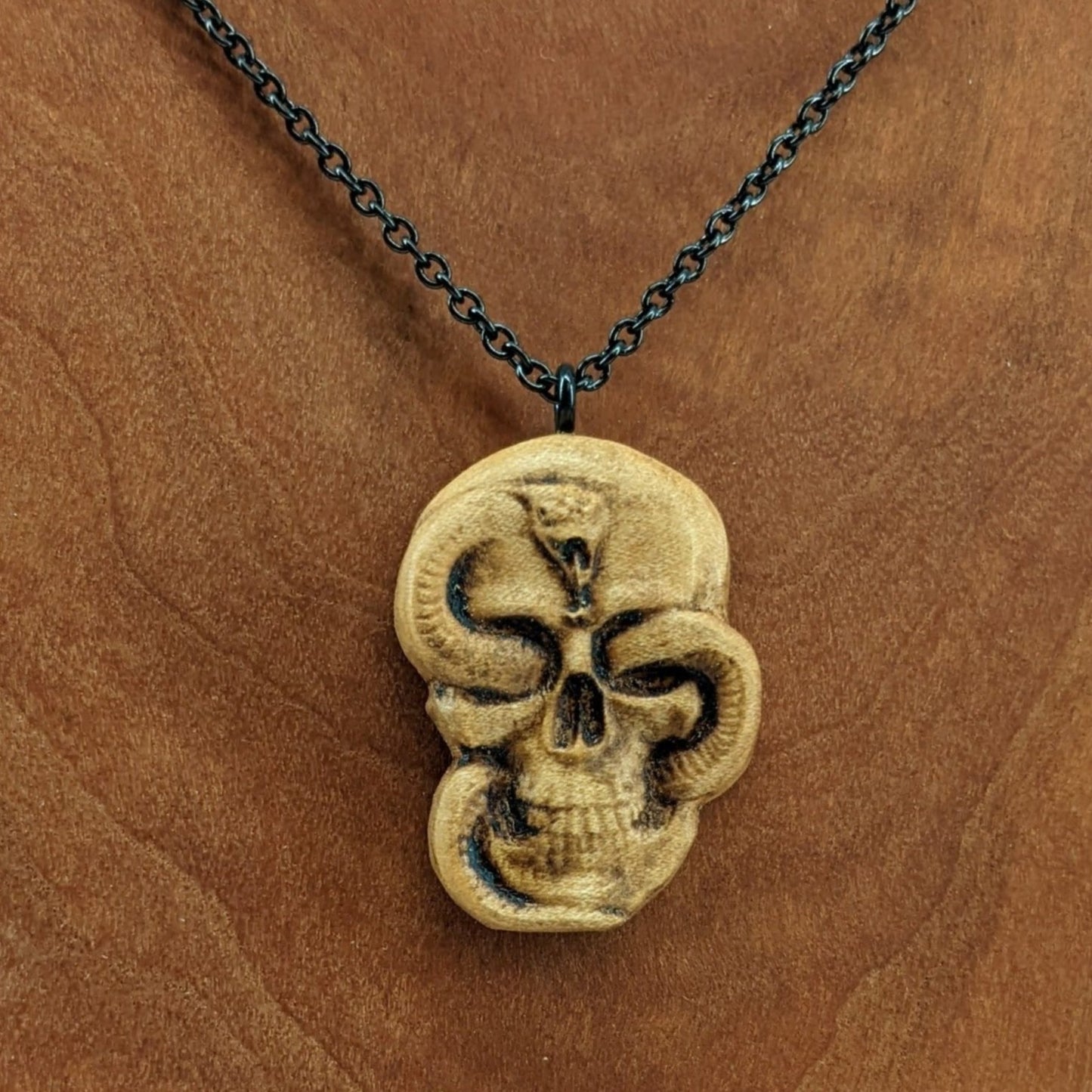 Wooden necklace pendant carved in the shape of a skull with a snake crawling through the eyes. Made from hard maple wood and hanging from a black stainless steel chain against a cherry wood display.