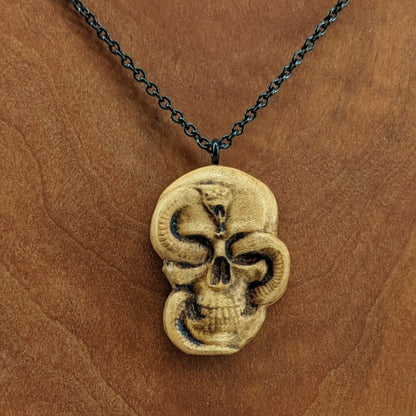 Wooden necklace pendant carved in the shape of a skull with a snake crawling through the eyes. Made from hard maple wood and hanging from a black stainless steel chain against a cherry wood display.