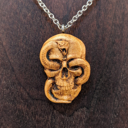 Wooden necklace pendant carved in the shape of a skull with a snake crawling through the eyes. Made from hard maple wood and hanging from a silver stainless steel chain against a dark walnut wood background.