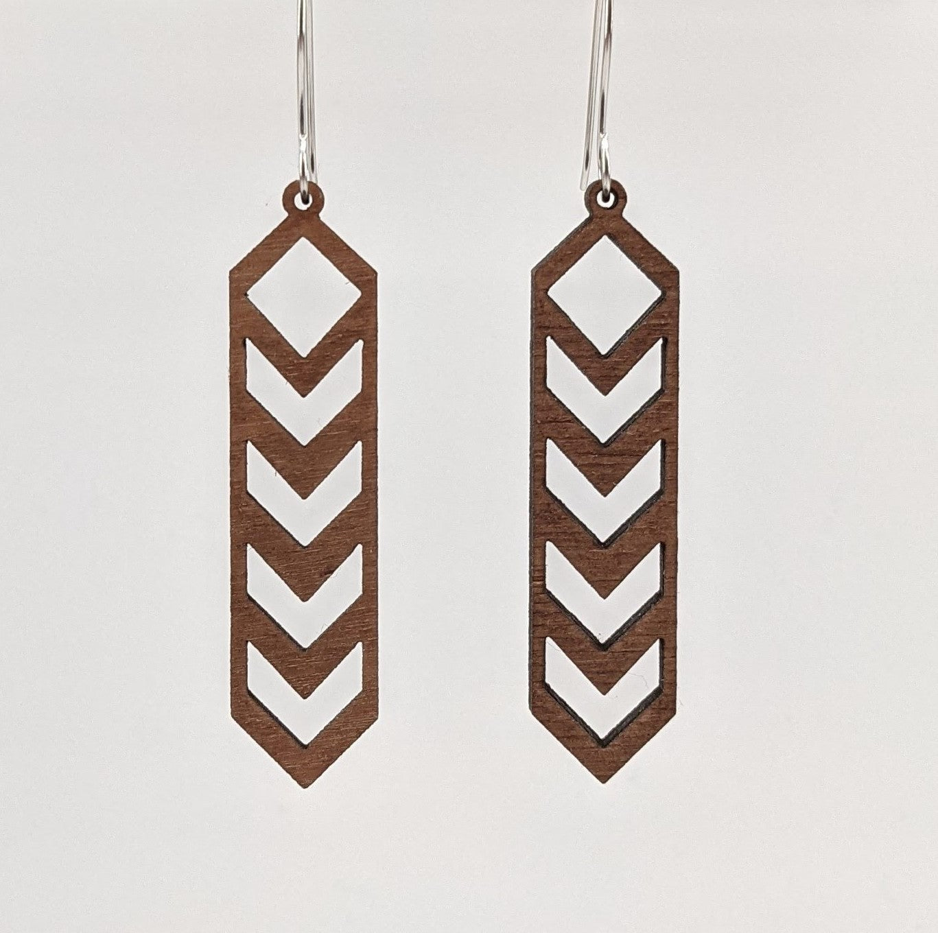 Pair of wooden earrings with silver stainless steel hooks. They are brown slim, elongated diamond shapes with downward chevron cutouts within. Made from birch wood hanging from a model Davidson Workshop sign against a white background.(Close up View)