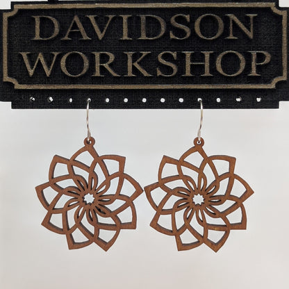 Pair of wooden earrings with silver stainless steel hooks. They are brown cutouts of flowers with spiraling, overlapping petals. Made from birch wood hanging from a model Davidson Workshop sign against a white background. (Close up View)