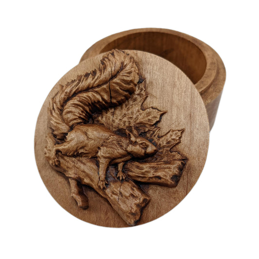 Round wooden box 3D carved with a squirrel with a large bushy tail and pointed ears standing on a forked  tree branch with maple leaves sticking out from behind. Made from hard maple wood stained brown against a white background.