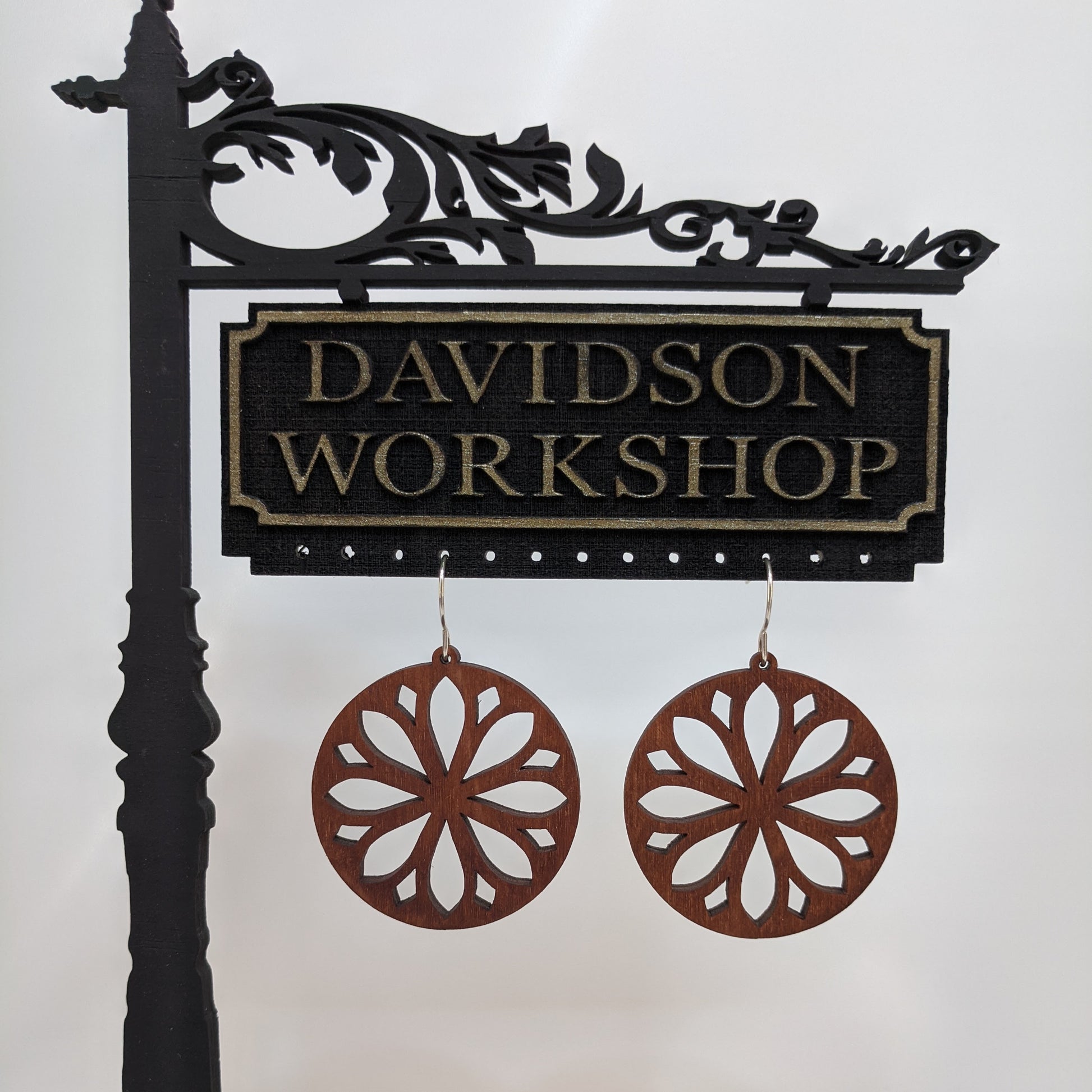 Pair of wooden earrings with silver stainless steel hooks. They are brown round with symmetrical petal shaped cutouts within. Made from birch wood hanging from a model Davidson Workshop sign against a white background. 