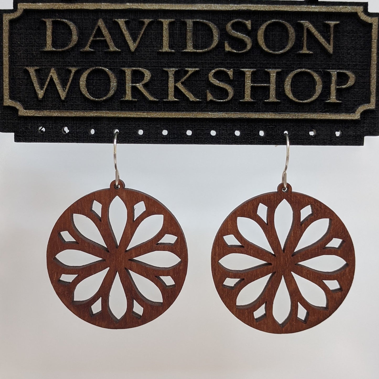 Pair of wooden earrings with silver stainless steel hooks. They are brown round with symmetrical petal shaped cutouts within. Made from birch wood hanging from a model Davidson Workshop sign against a white background. (Close up View)