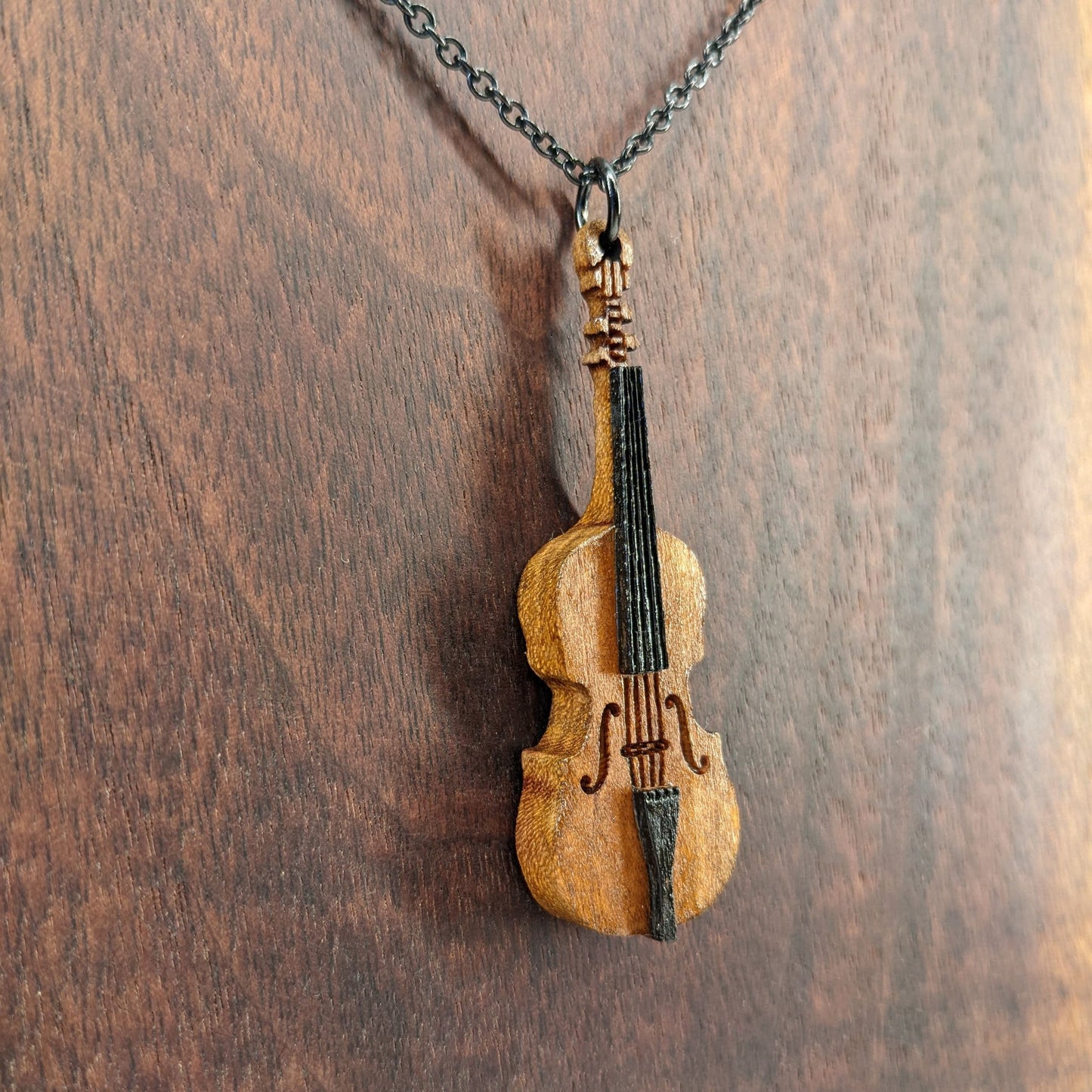 Wooden necklace pendant created in the shape of a miniature cello stand up bass. Made from cherry wood and hanging from a black stainless steel chain against a dark walnut background.