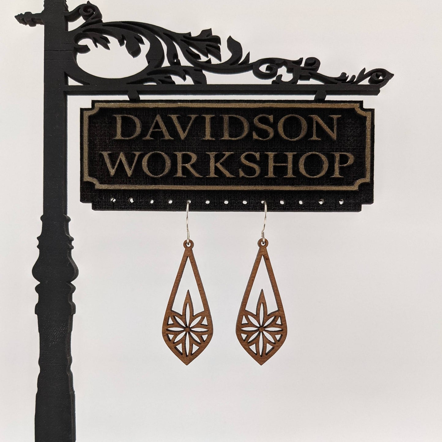 Pair of wooden earrings with silver stainless steel hooks. They are long brown teardrops with an 8 pointed star with sharp points within. Made from birch wood hanging from a model Davidson Workshop sign against a white background.