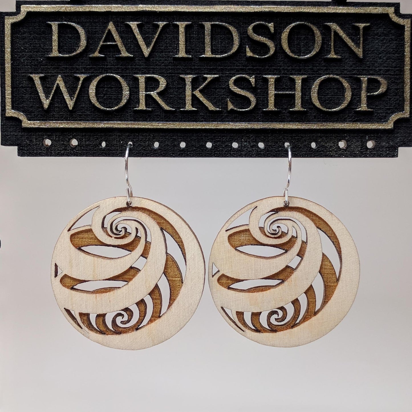 Pair of wooden earrings with silver stainless steel hooks. They are light finish balls with spirals engraved creating a 3D spherical illusion. Made from birch wood hanging from a model Davidson Workshop sign against a white background.(Close up View)