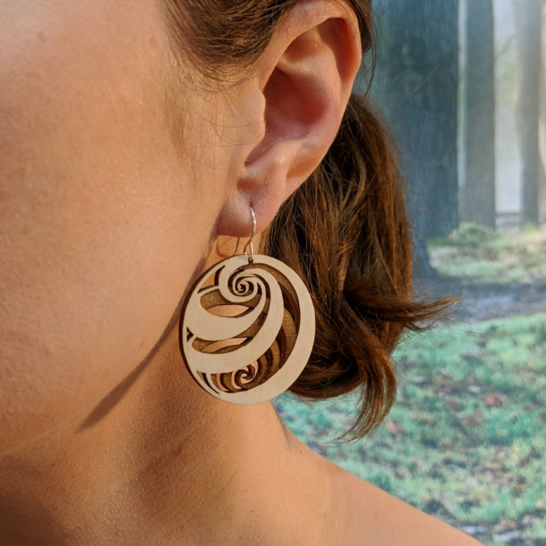 Pair of wooden earrings with silver stainless steel hooks. They are light finish balls with spirals engraved creating a 3D spherical illusion. Made from birch wood, being modeled by a women in front of a nature scene.