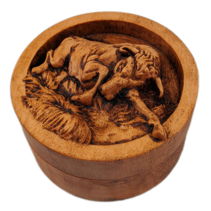 Round wooden box 3D carved with a angry bull charging forward stomping over small grassy patches on the ground. Its head is angled downward with its curved horns pointed ahead. Made from hard maple wood stained brown against a white background.