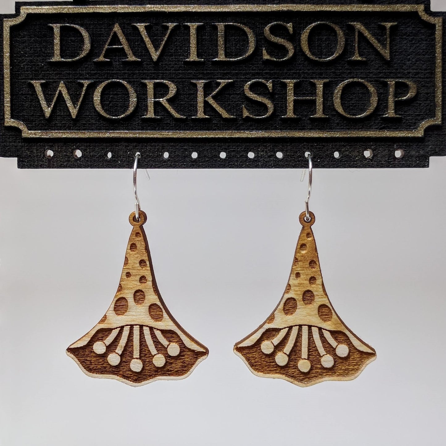 Pair of wooden earrings with silver stainless steel hooks. They are natural finish polka dotted petals with multiple pistols in their center. Made from birch wood hanging from a model Davidson Workshop sign against a white background. (Close up View)