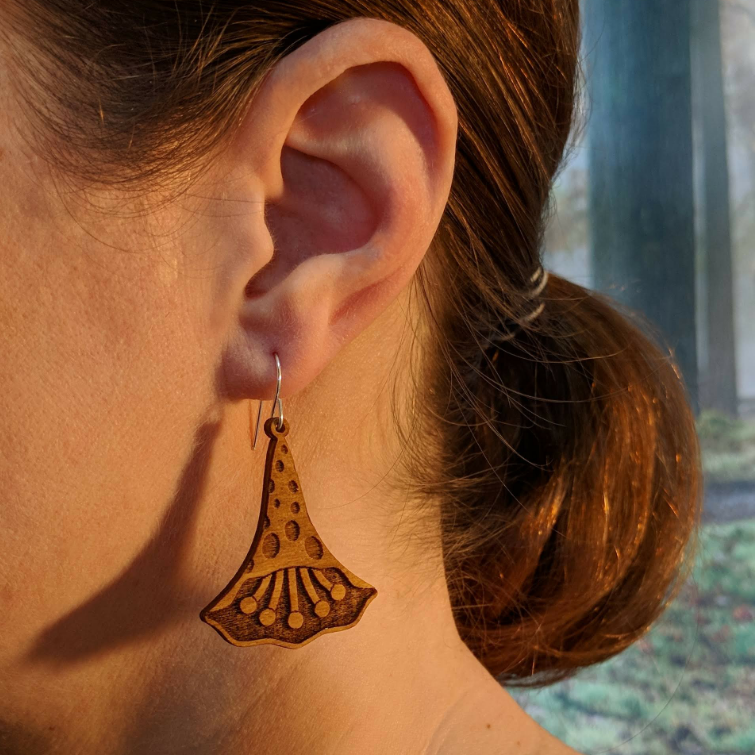 Pair of wooden earrings with silver stainless steel hooks. They are natural finish polka dotted petals with multiple pistols in their center. Made from birch wood, being modeled by a women in front of a nature scene.