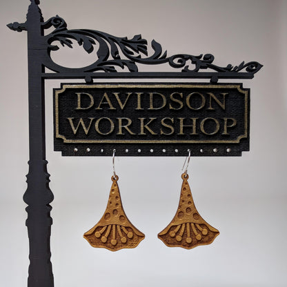 Pair of wooden earrings with silver stainless steel hooks. They are natural finish polka dotted petals with multiple pistols in their center. Made from birch wood hanging from a model Davidson Workshop sign against a white background. 