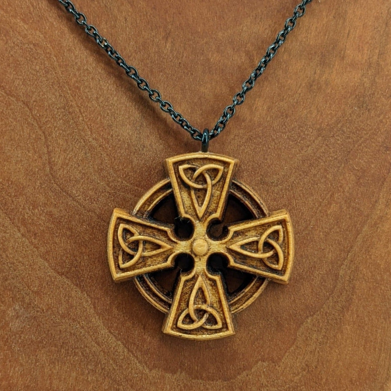 Wooden necklace pendant created in the shape of a Celtic cross. Made from hard maple wood and hanging from a black stainless steel chain against a cherry wood background.