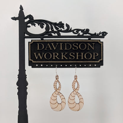 Pair of wooden earrings with silver stainless steel hooks. They are light natural finish scaled infinity symbols twisting over themselves.  Made from birch wood hanging from a model Davidson Workshop sign against a white background.