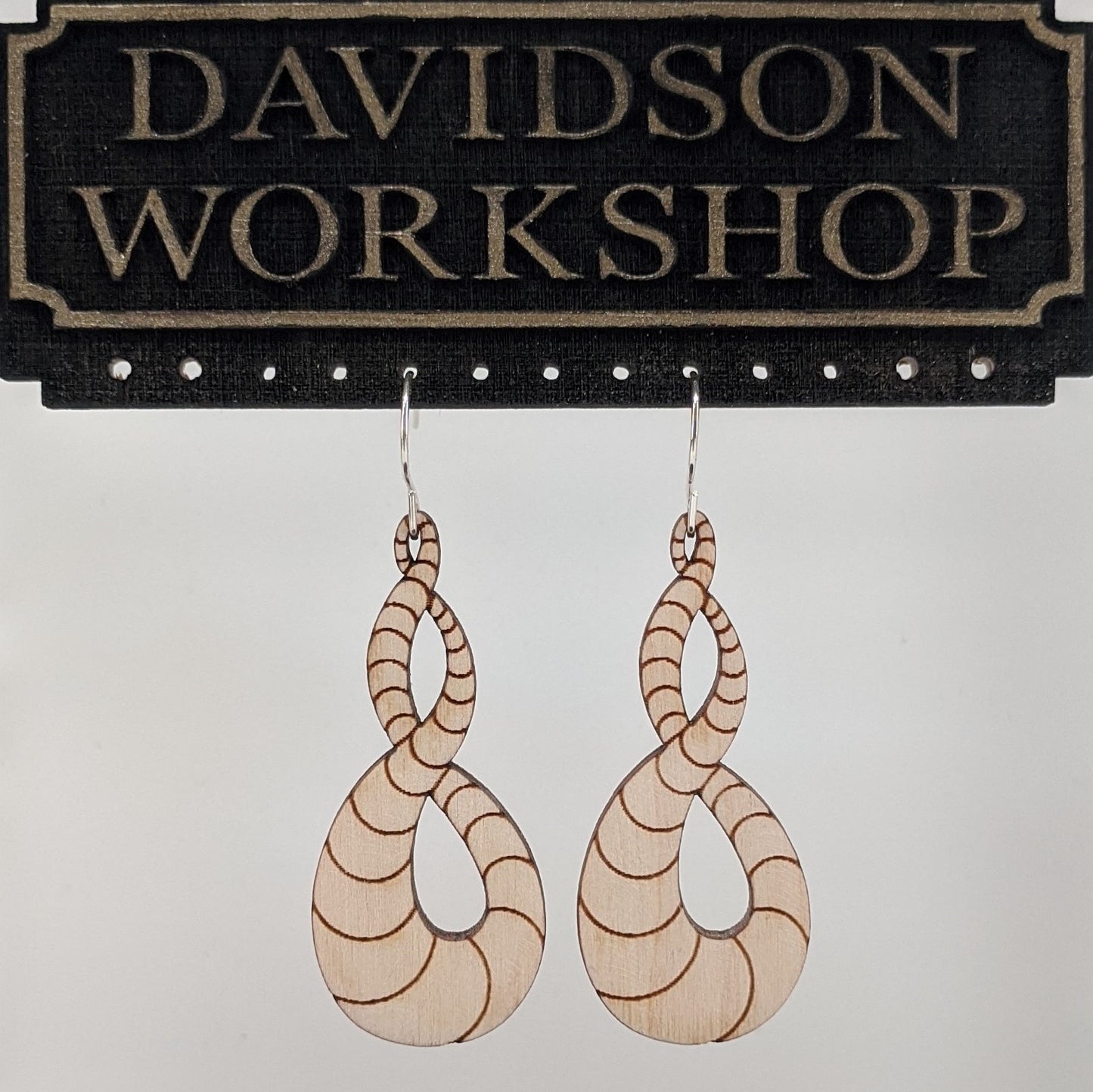 Pair of wooden earrings with silver stainless steel hooks. They are light natural finish scaled infinity symbols twisting over themselves.  Made from birch wood hanging from a model Davidson Workshop sign against a white background. (Close up View)