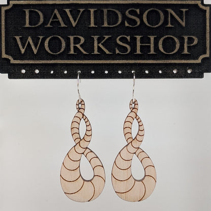 Pair of wooden earrings with silver stainless steel hooks. They are light natural finish scaled infinity symbols twisting over themselves.  Made from birch wood hanging from a model Davidson Workshop sign against a white background. (Close up View)