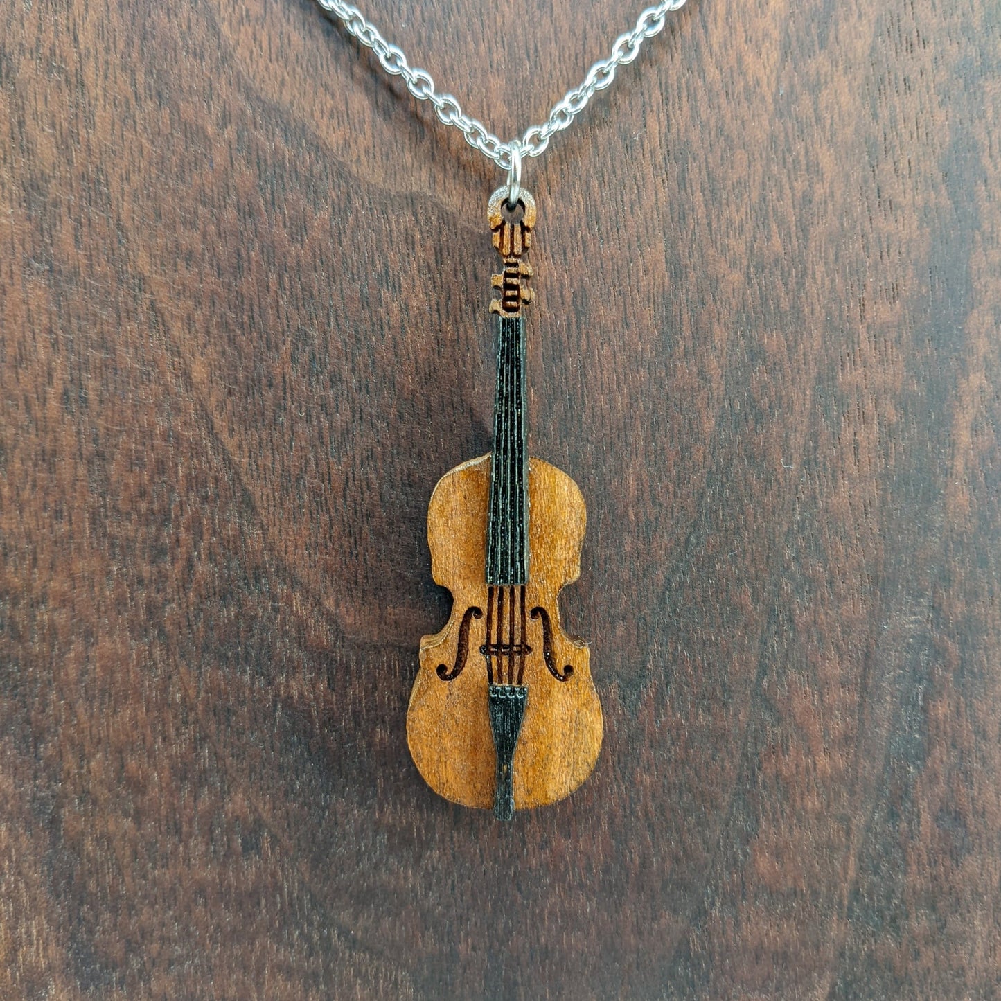 Wooden necklace pendant created in the shape of a miniature cello stand up bass. Made from cherry wood and hanging from a silver stainless steel chain against a dark walnut background.