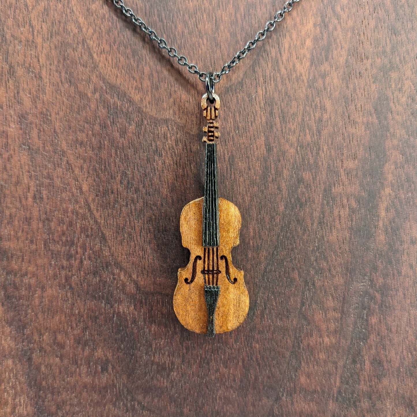 Wooden necklace pendant created in the shape of a miniature cello stand up bass. Made from cherry wood and hanging from a black stainless steel chain against a dark walnut background.