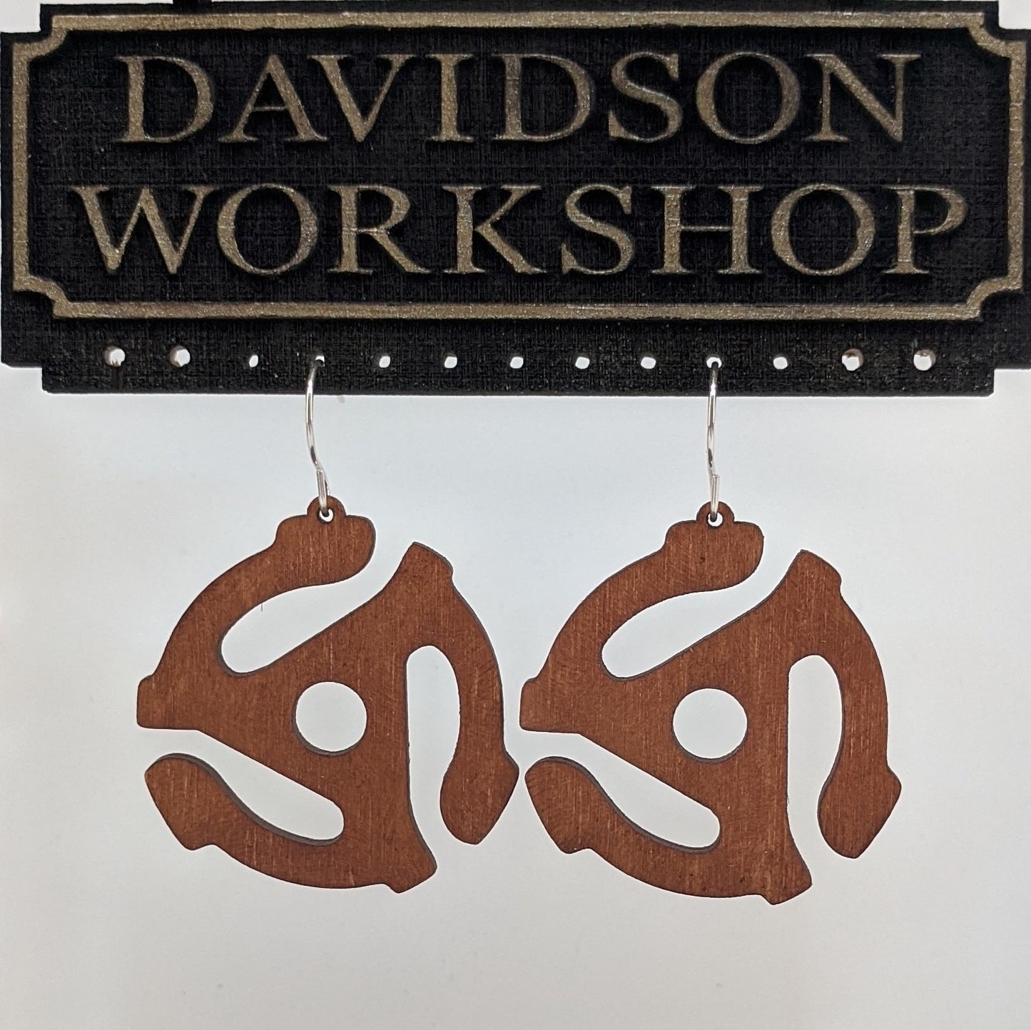 Pair of wooden earrings with silver stainless steel hooks. They are brown 3 armed spiraling models of vintage 45mm record player adapters. Made from birch wood hanging from a model Davidson Workshop sign against a white background. (Close up View)