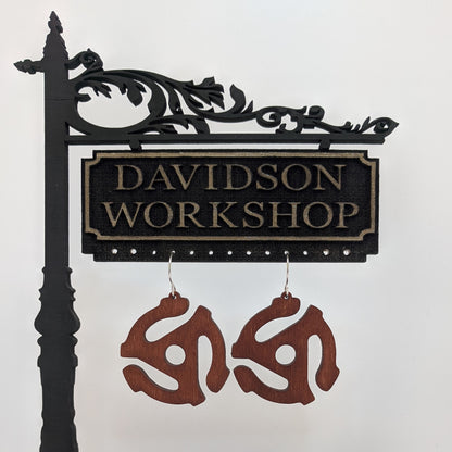 Pair of wooden earrings with silver stainless steel hooks. They are brown 3 armed spiraling models of vintage 45mm record player adapters. Made from birch wood hanging from a model Davidson Workshop sign against a white background.