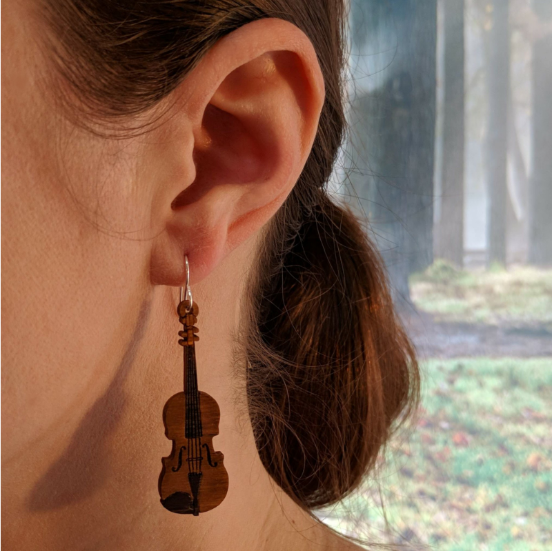 Pair of wooden earrings with silver stainless steel hooks. They are brown violins with engraved detailing and hand painted accents. Made from birch wood, being modeled by a women in front of a nature scene.